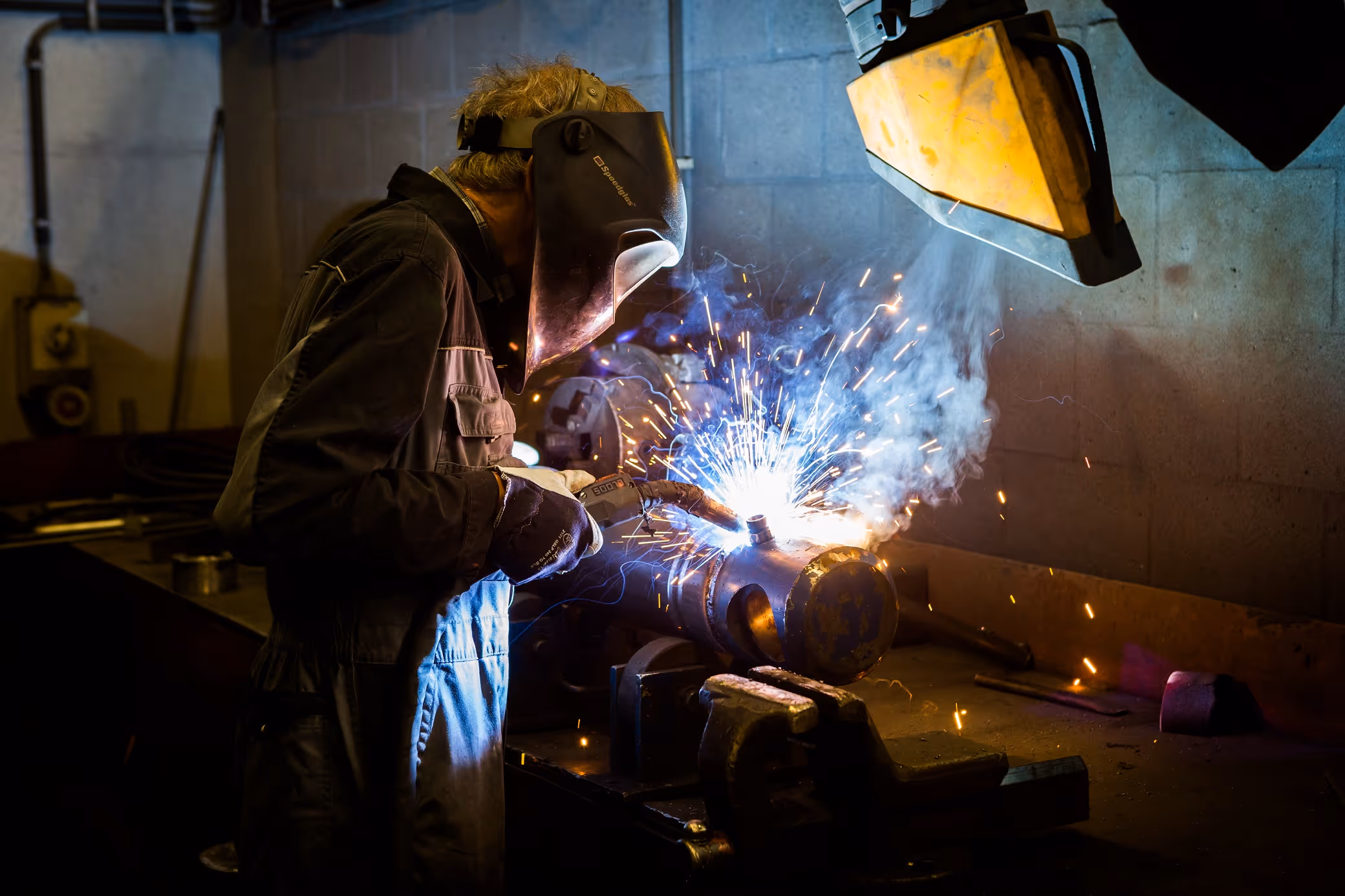 Lasser met beschermhelm en handschoenen bezig met lassen van een metalen onderdeel op werkbank.