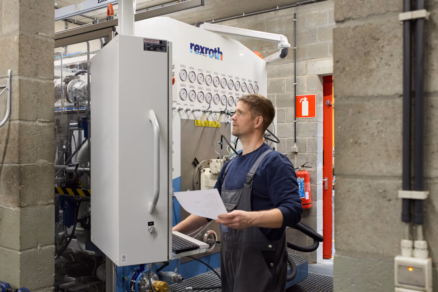 Man in werkoveralls bedient een industriële machine met drukmeters terwijl hij een document vasthoudt.
