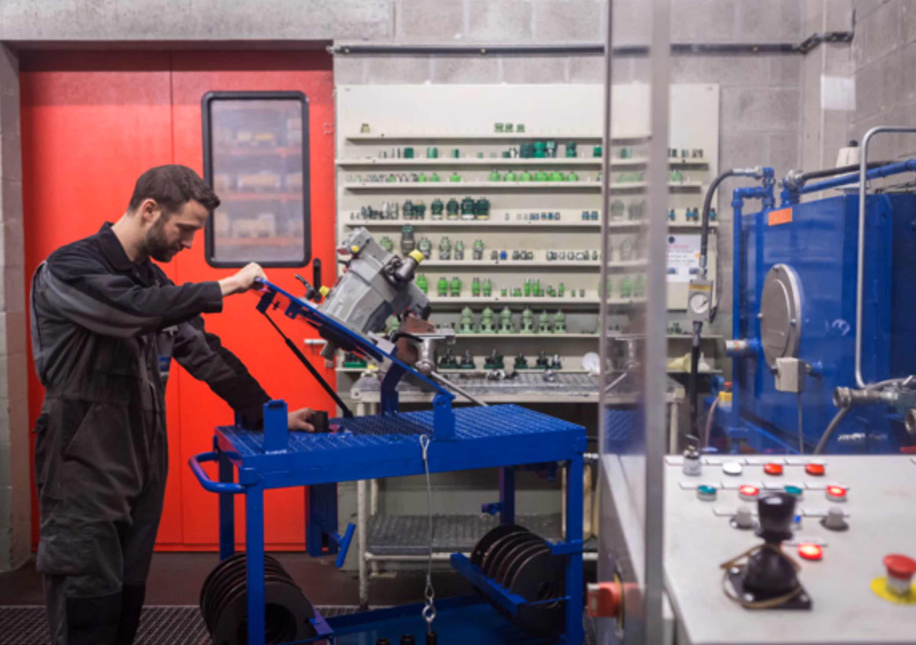 Een technicus in donker werkkleding werkt met apparatuur op een blauwe werktafel in een industrieel laboratorium met betonnen muren en een rode deur achter hem.