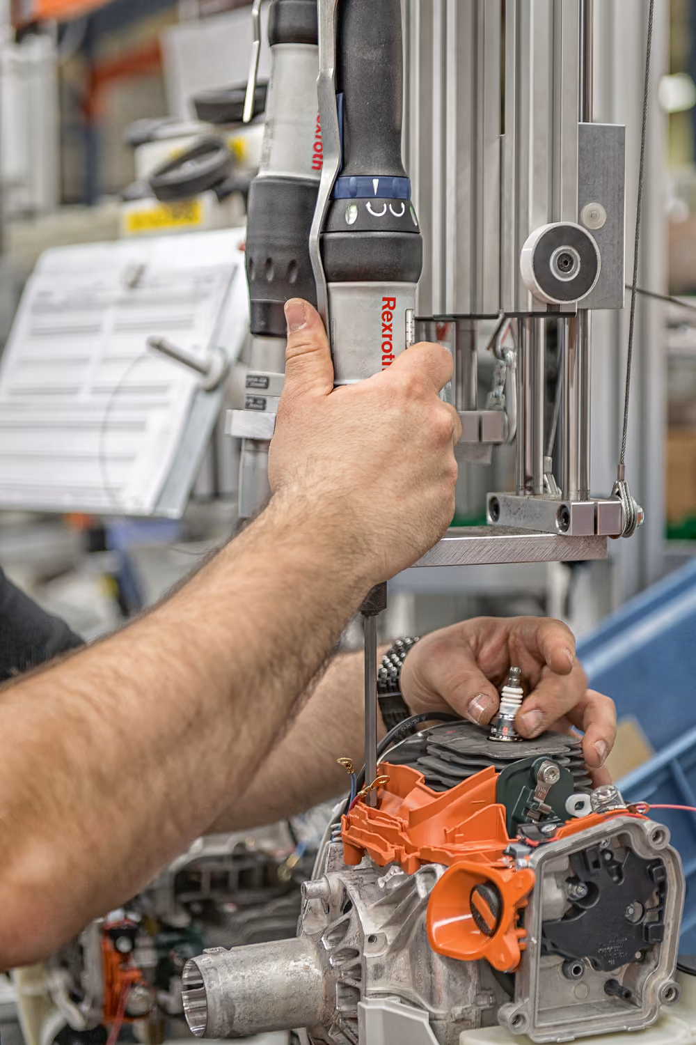 Een persoon gebruikt een elektrisch gereedschap met het merk Rexroth om een mechanisch onderdeel met oranje en zilveren delen te monteren of repareren, waarbij een bougie wordt vastgehouden.