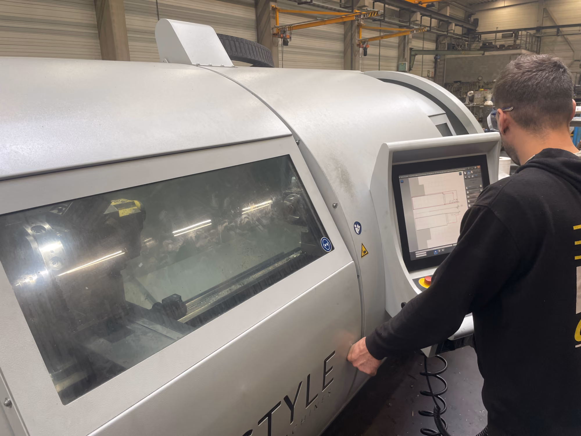 Een man staat in een fabriekshal voor een CNC-machine en bedient het touchscreen aan de zijkant van de machine.
