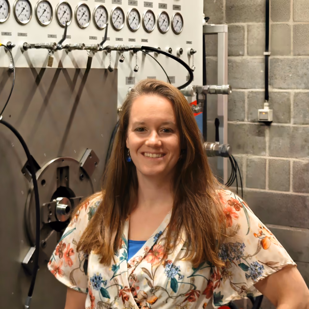 Vrouw met lang bruin haar en een bloemrijke blouse staat glimlachend voor een industrieel bedieningspaneel met meerdere manometers en kleppen.