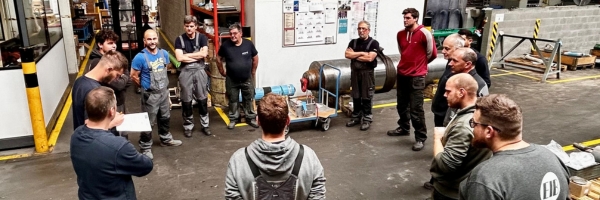 Een groep van elf mannen staat in een cirkel in een fabriekshal en luistert naar een man die een paper in zijn hand houdt en spreekt.
