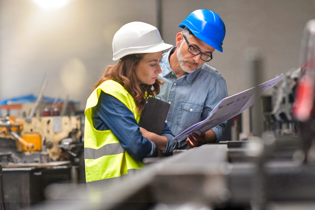 An older man and a younger woman, both wearing hard hats, look over paperwork together at a factory. 