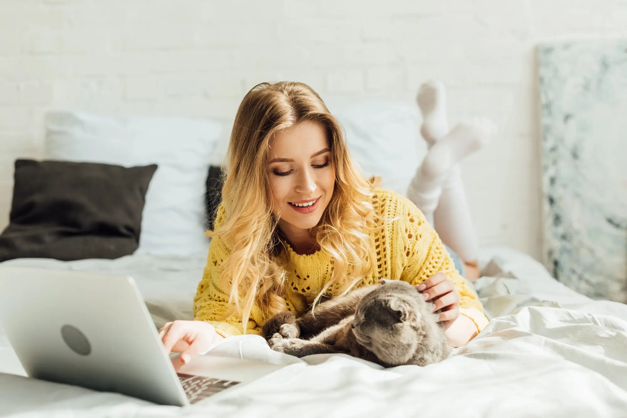 Woman in yellow sweater using laptop with cat on bed.