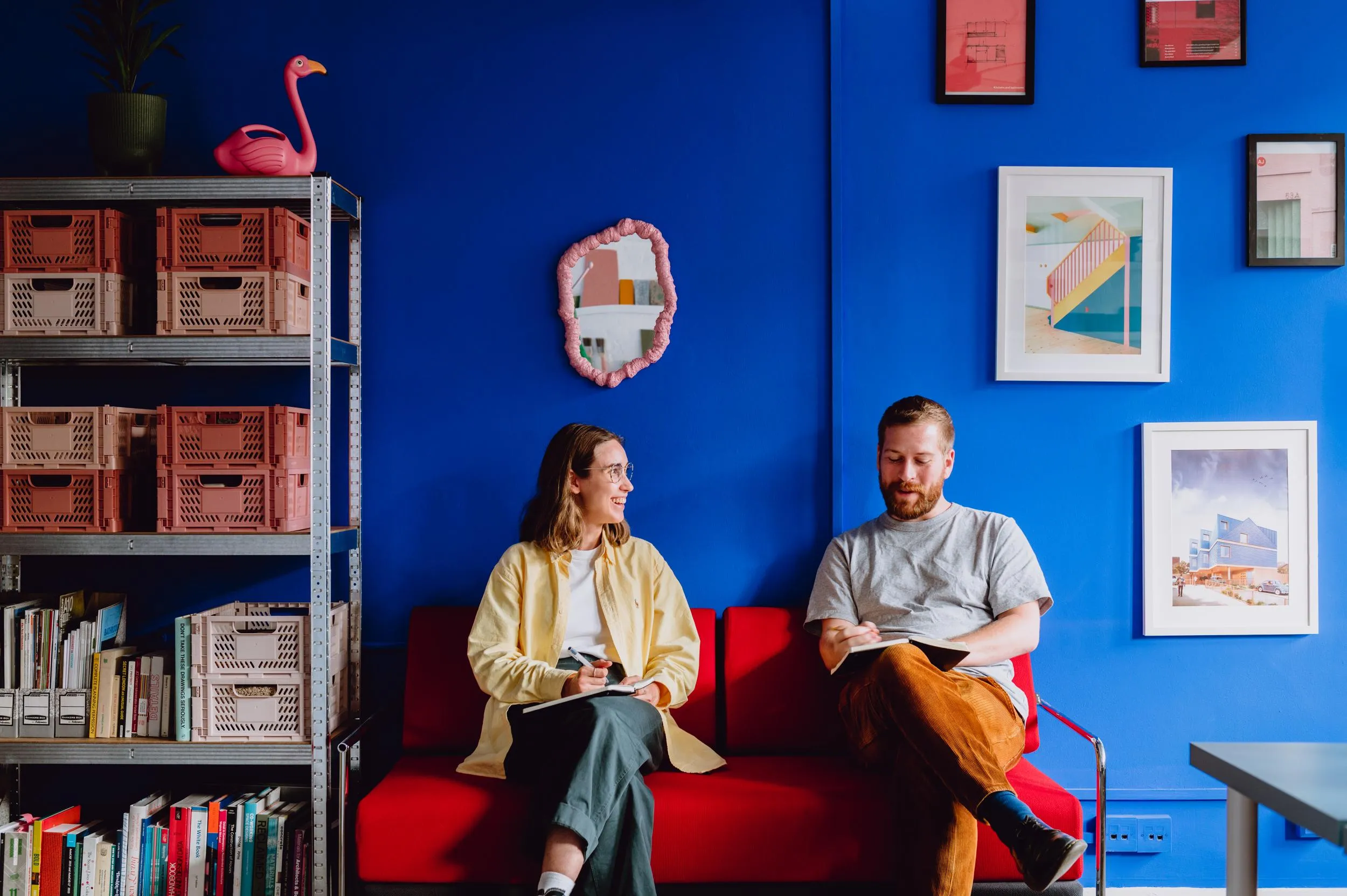 Photo of two team members sat on the red sofa, with a shelving unit to the side and blue accent wall in the background. By Office S&M, RIBA Chartered Architectural Practice in London.