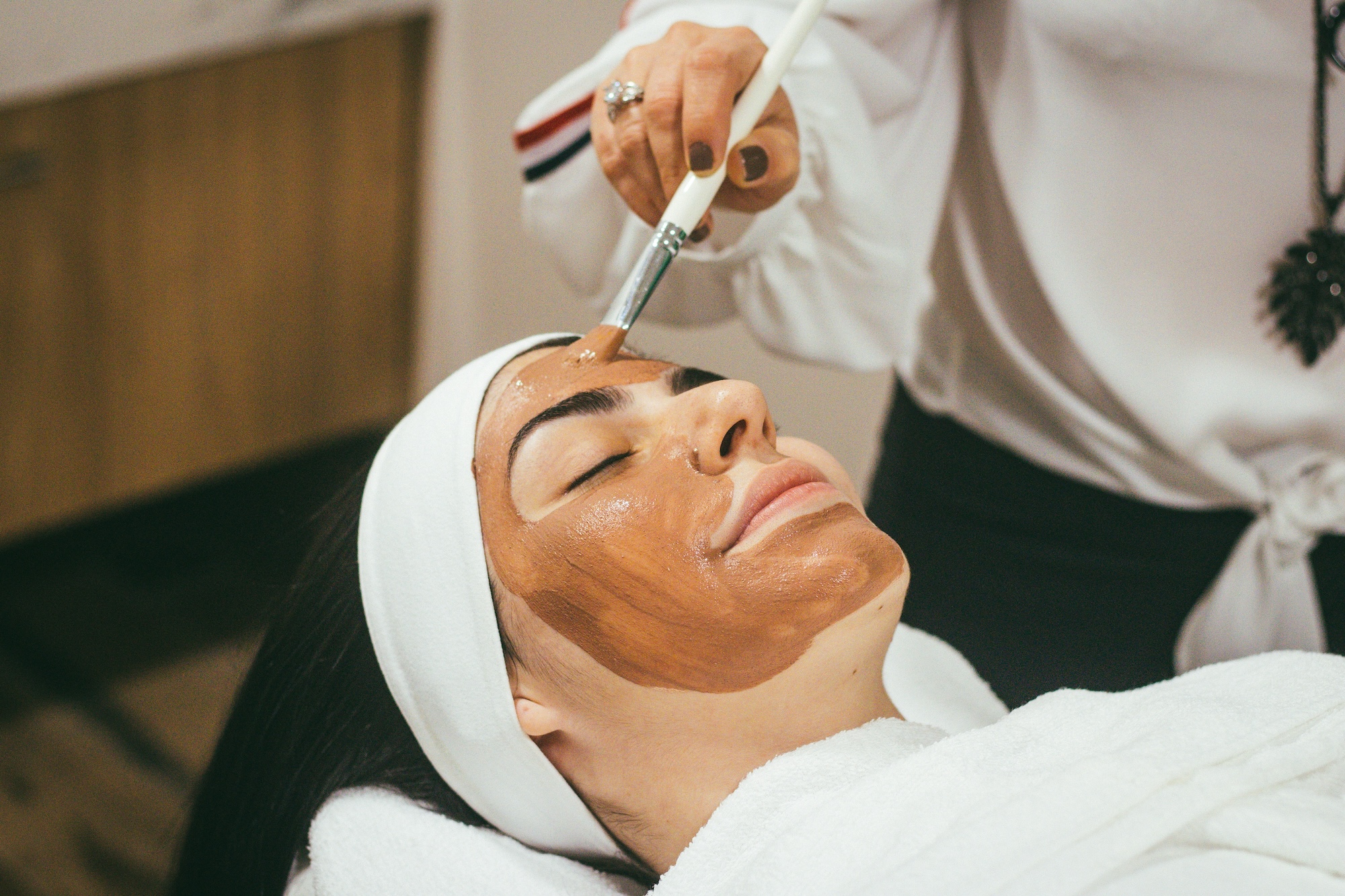 A woman lays on a spa bed witha placid look while a technician applies a mask to her face.
