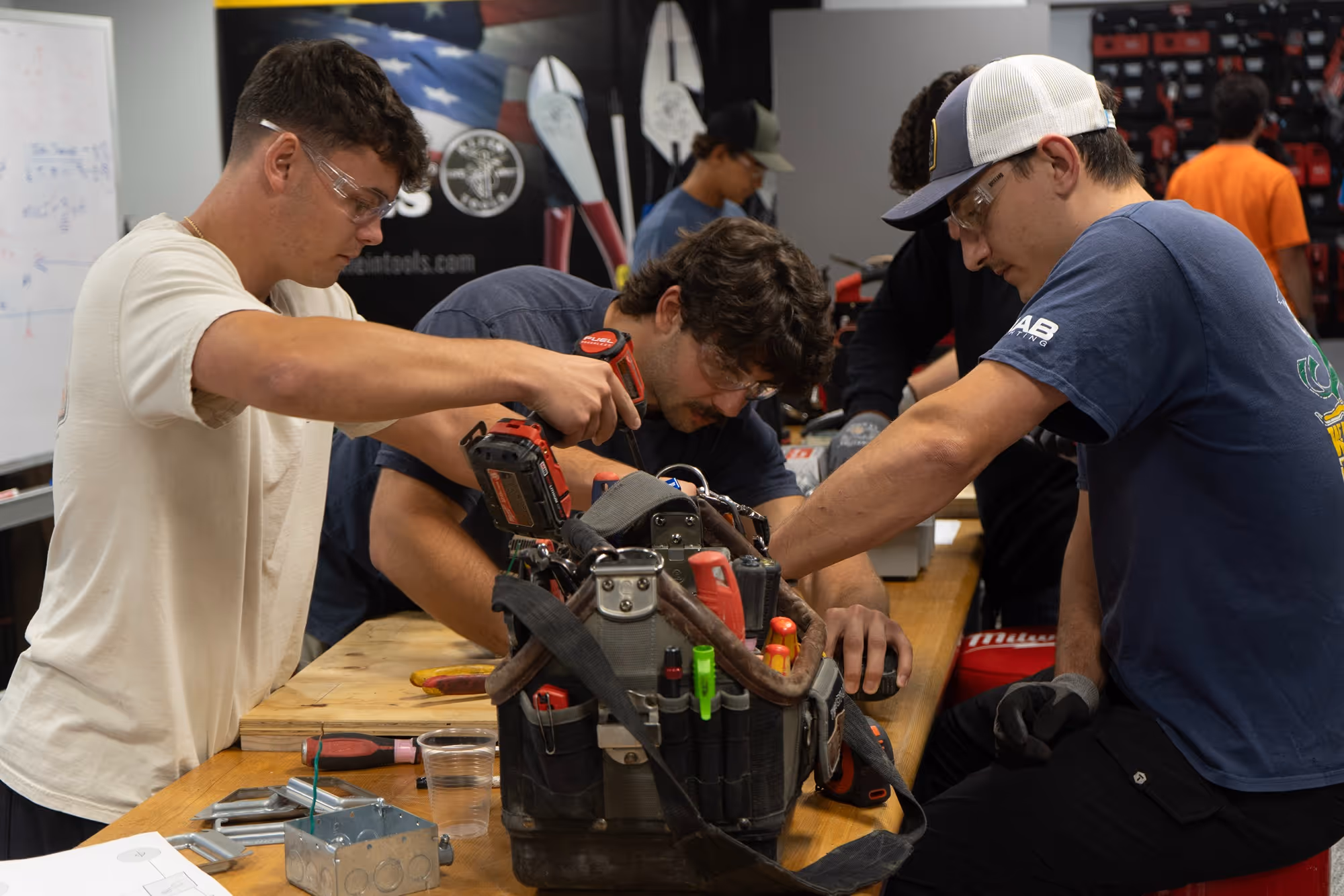 Three young men wearing safety glasses work together on a mechanical project at a wooden table with tools and equipment.