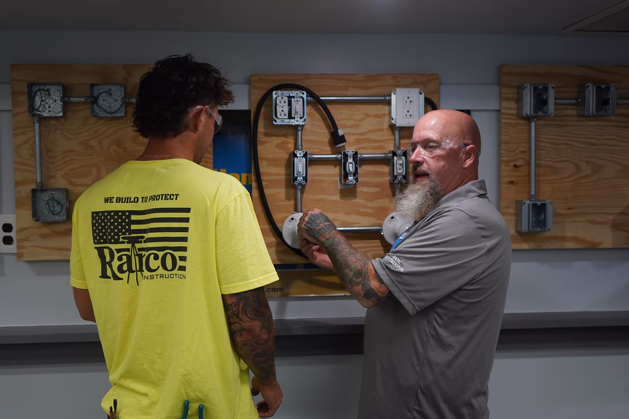 Two men in safety glasses discussing electrical wiring on mounted wooden boards with outlets and switches.