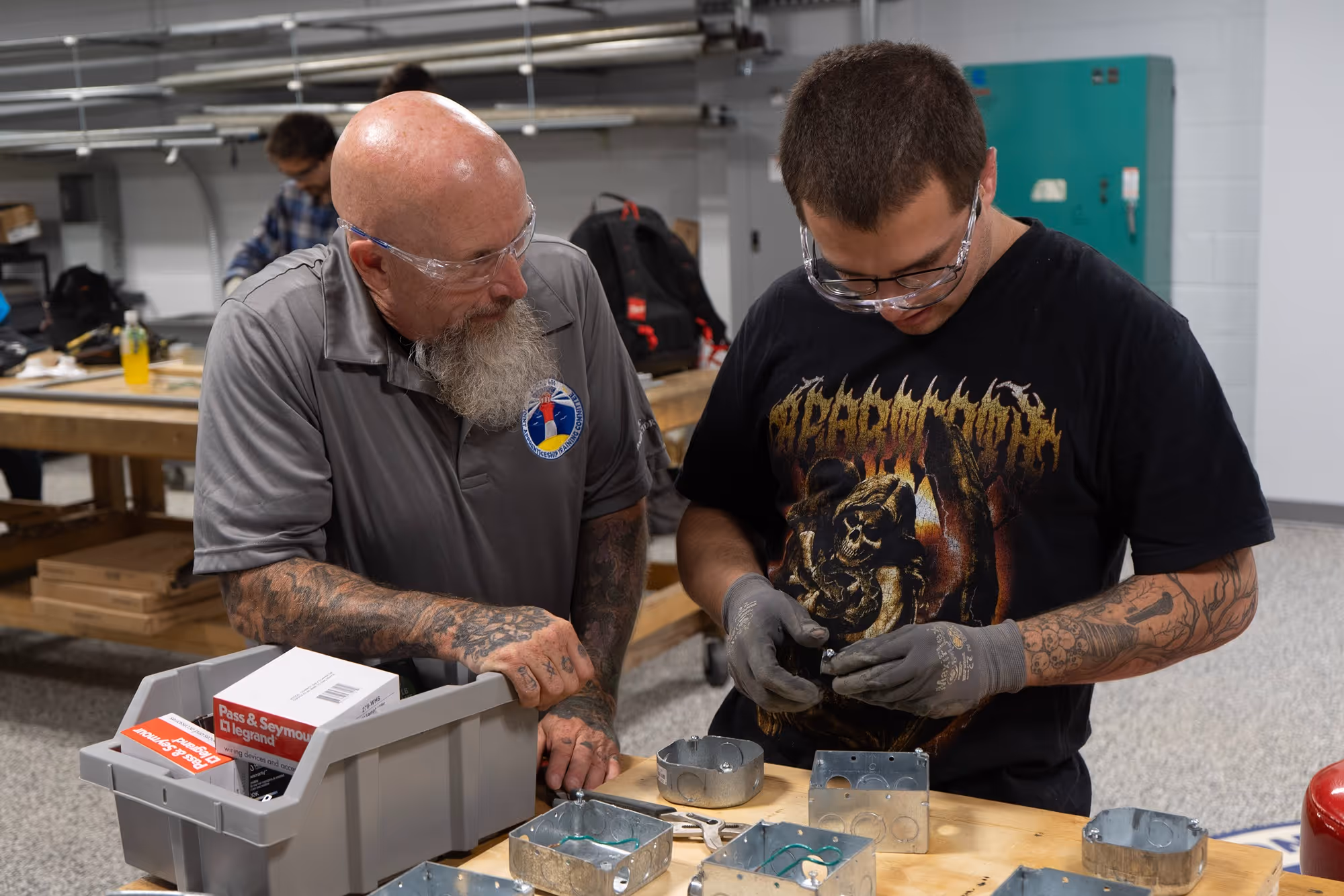 Two men wearing safety glasses working on electrical wiring with metal electrical boxes on a workbench in a workshop.