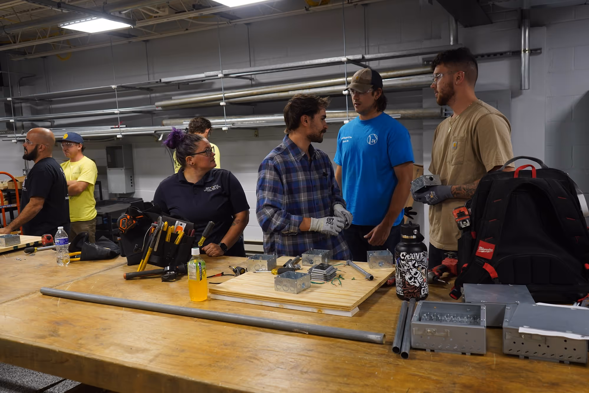 Group of people in a workshop discussing around a table with electrical boxes, tools, and metal pipes.