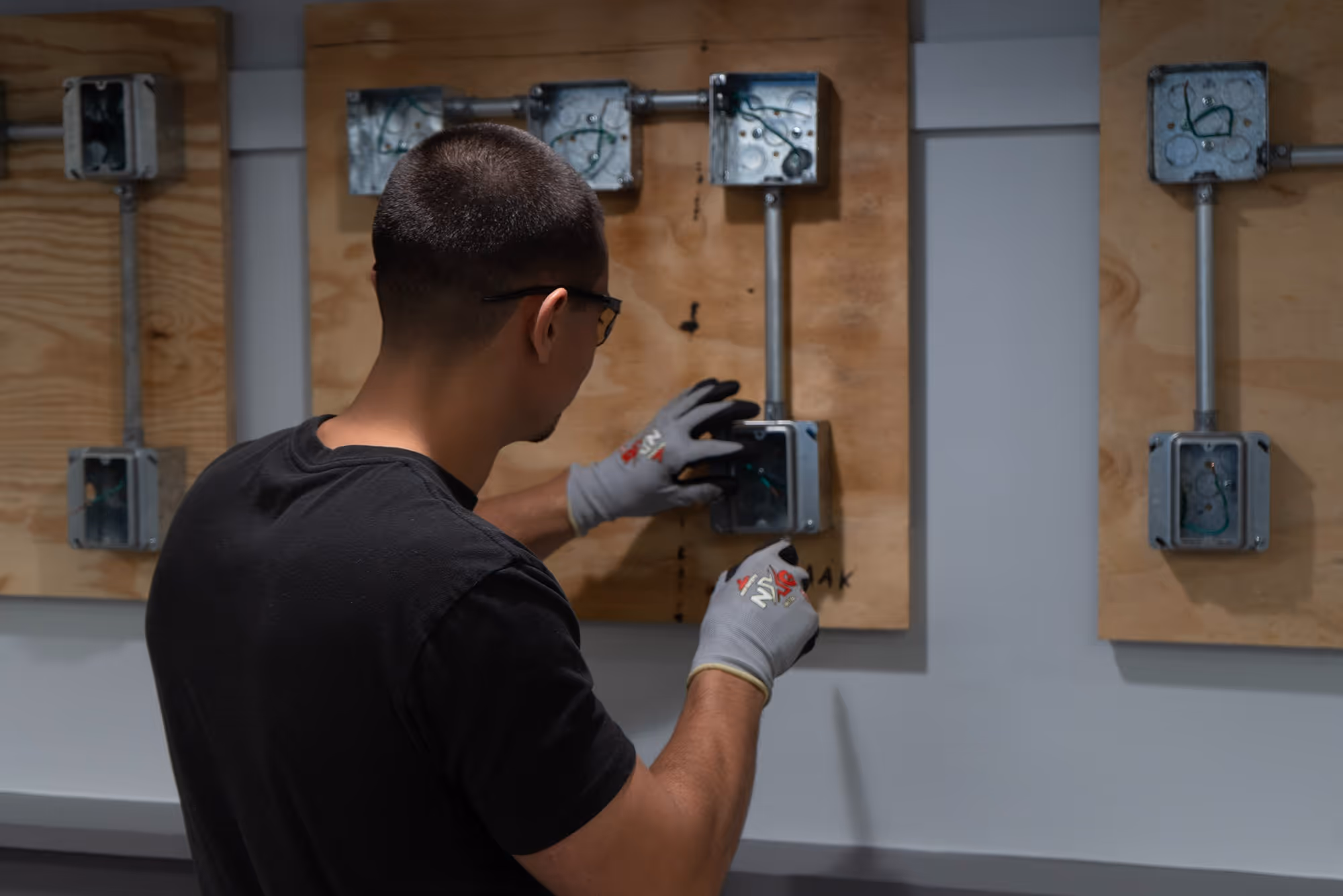 Three young men wearing safety glasses work together on a mechanical project at a wooden table with tools and equipment.