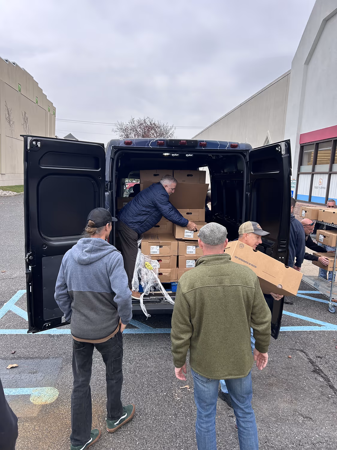 Men unloading cardboard boxes from the back of a black van parked in a lot.