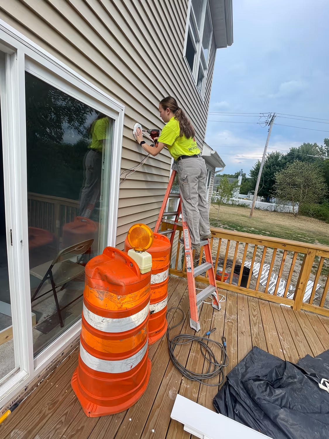 Person on a ladder installing or working on an electrical fixture on the exterior wall of a house with two orange traffic barrels nearby.