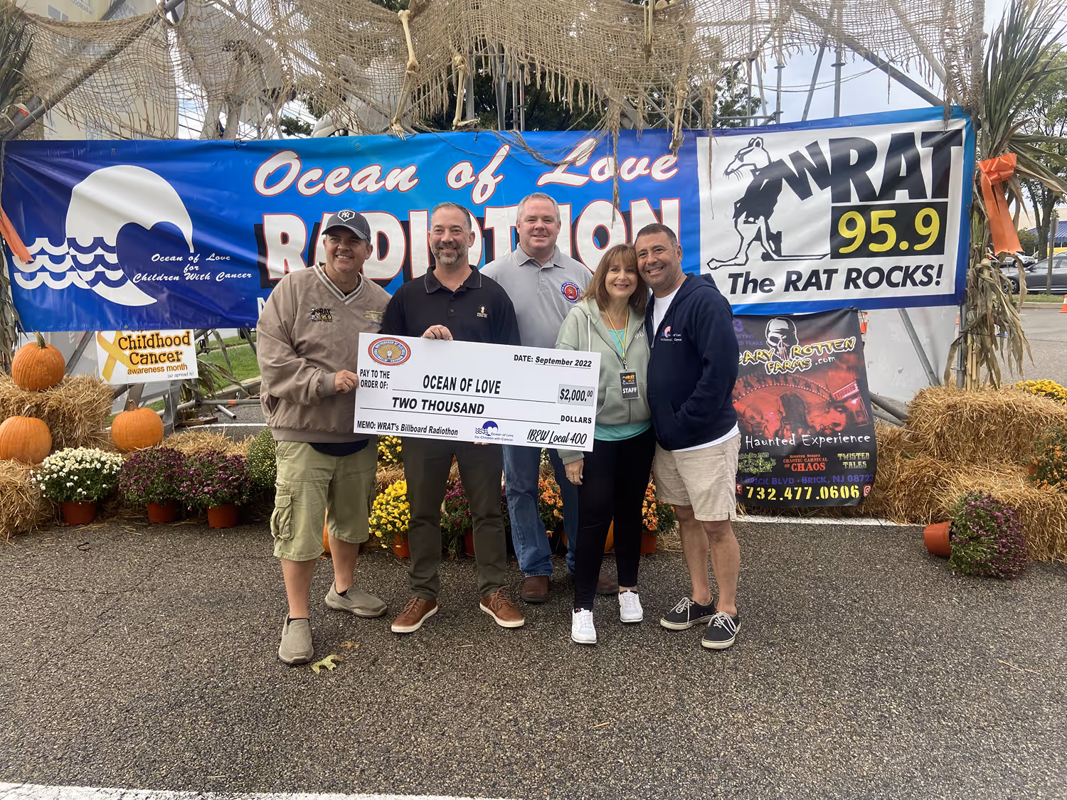 Five people standing in front of banners holding a large $2,000 check for Ocean of Love charity, with pumpkins and flowers nearby.
