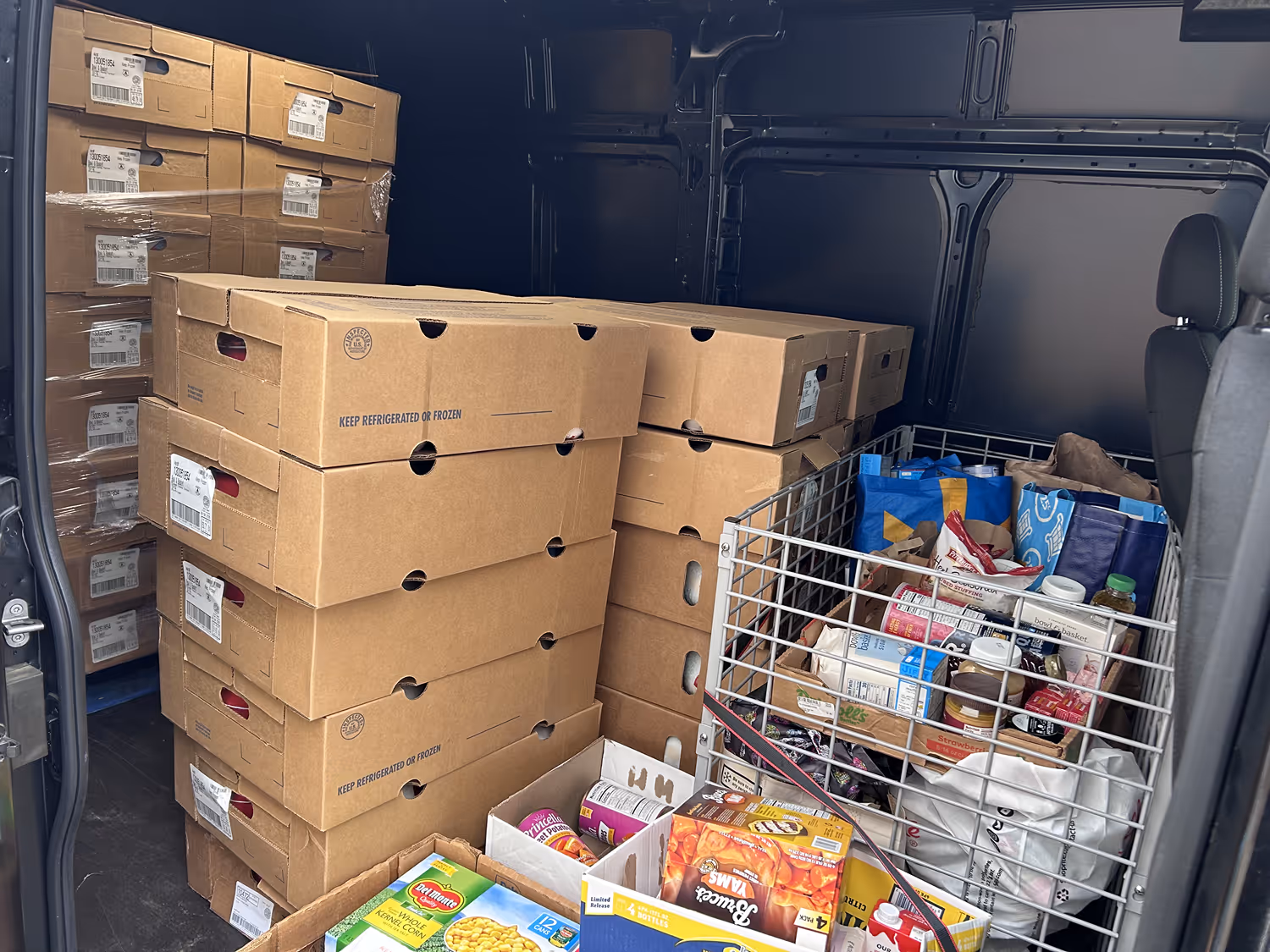 Interior of a delivery van loaded with stacked cardboard boxes and a metal shopping cart filled with grocery bags and food items.