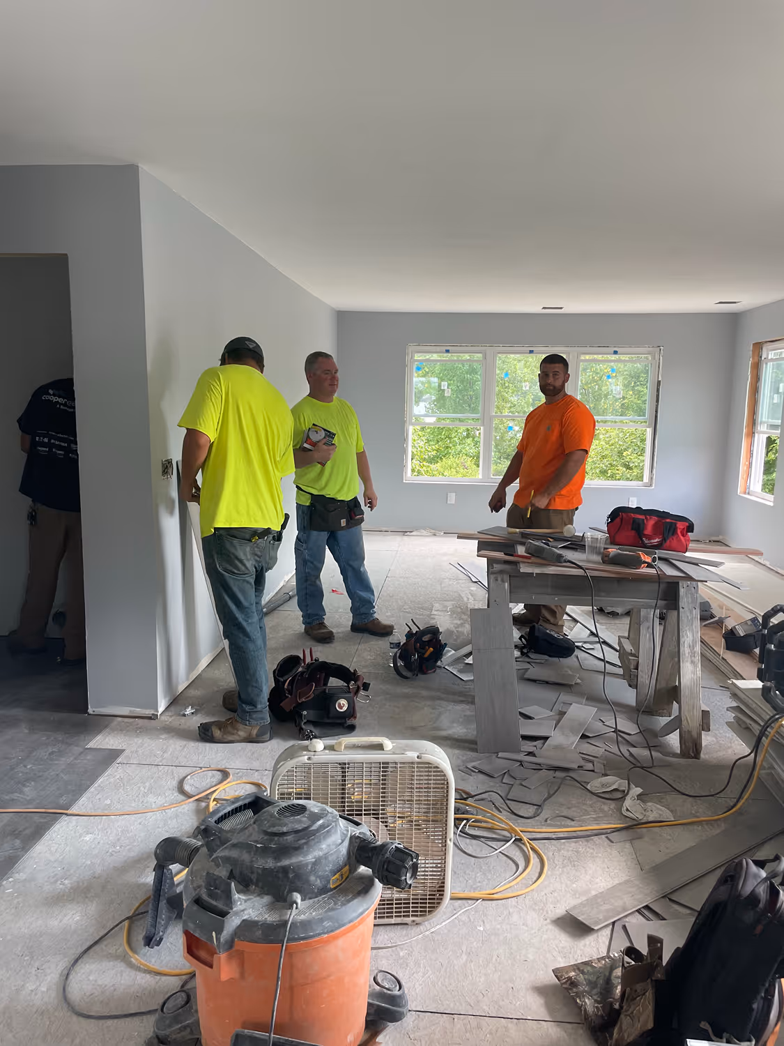 Three construction workers standing in an unfinished room with tools, tile pieces, and equipment scattered on the floor.