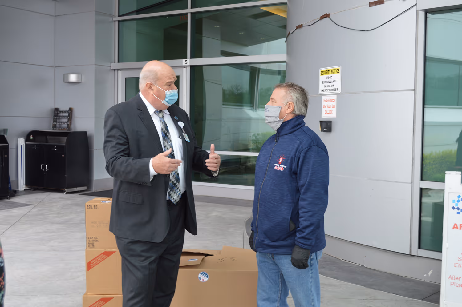 Two men wearing face masks talking outside a building near stacked cardboard boxes.