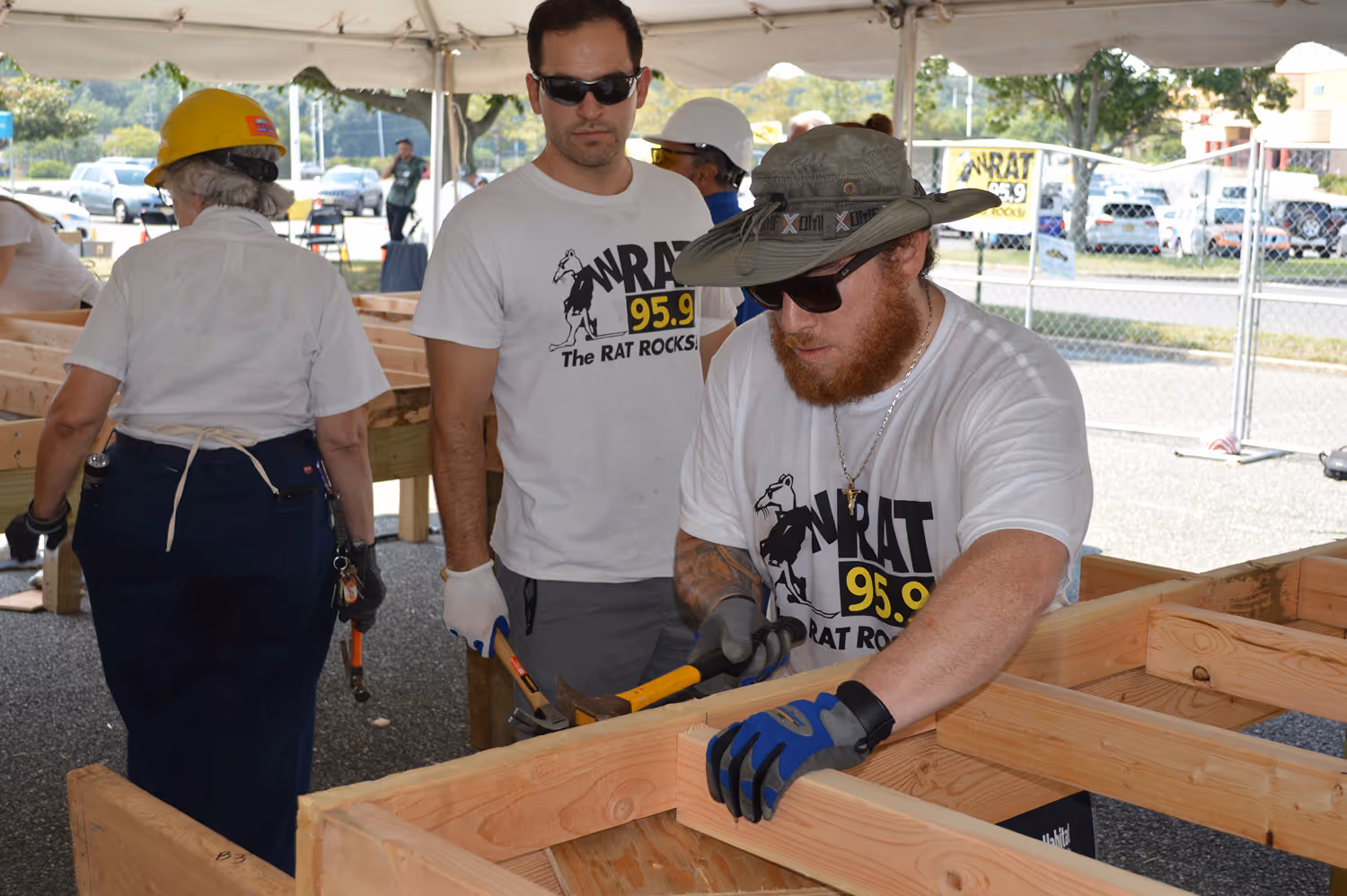 Two men wearing 'The RAT ROCKS 95.9' t-shirts working on wooden framing under a tent, one hammering a nail and the other holding tools, with a woman in a yellow hard hat nearby.