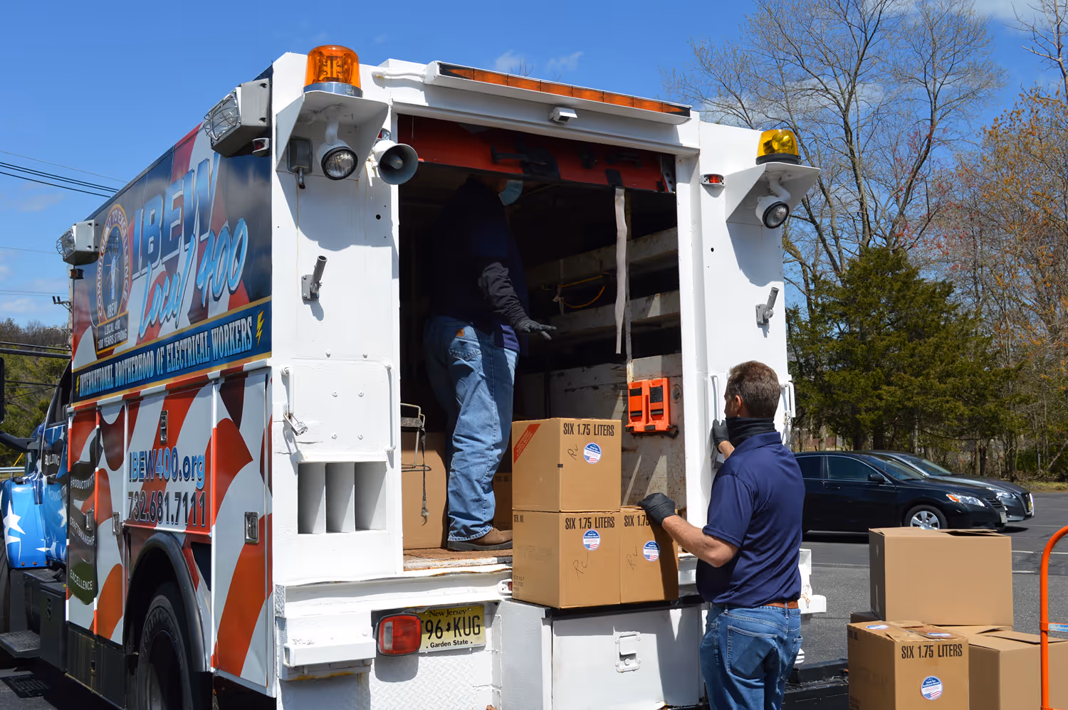 Group of people in a kitchen with trays of sandwiches and side dishes on a counter, some wearing blue shirts and hoodies.