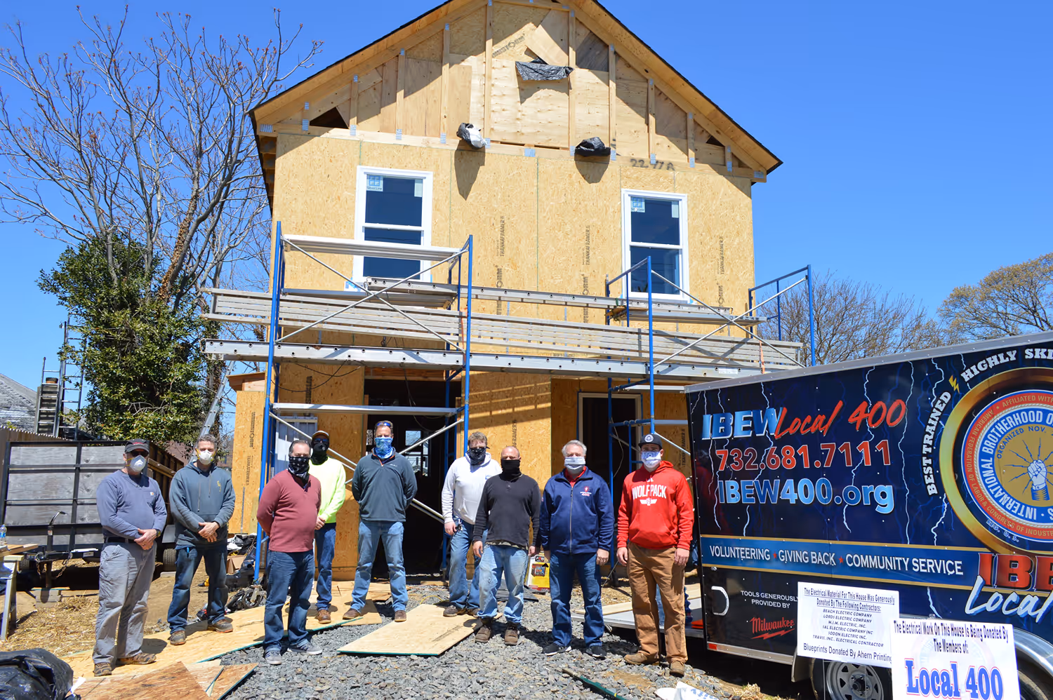 Group of nine construction workers posing in front of a partially built house with green sheathing and black brick foundation under a blue sky.