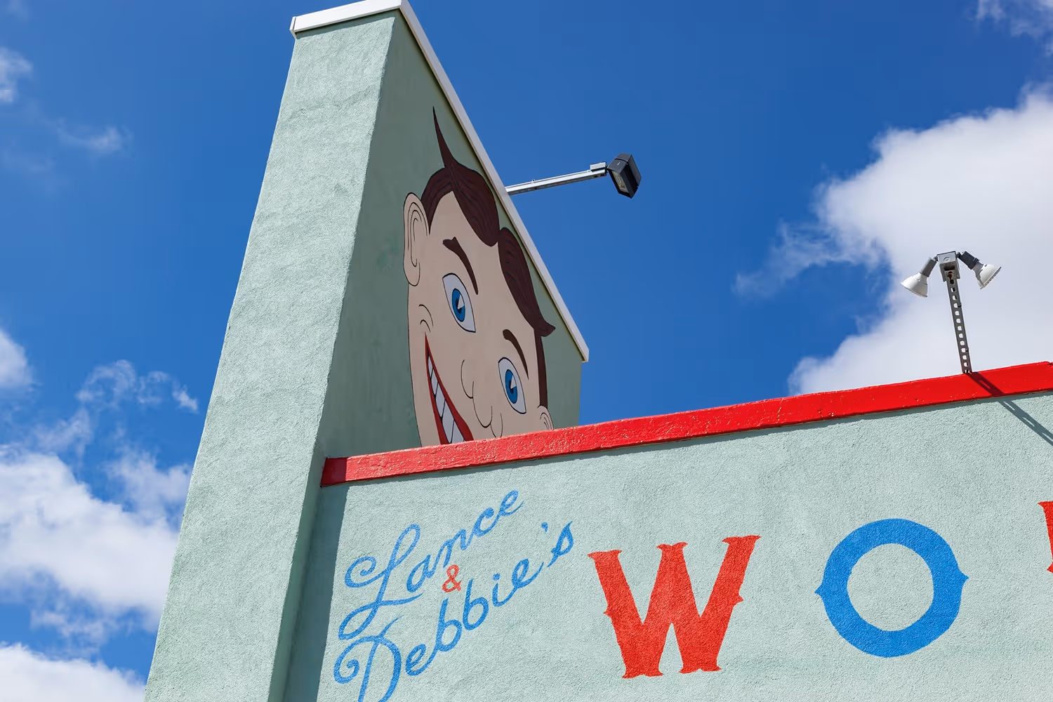 Murals on a light green building wall with the text 'Lance & Debbie's' in blue script and a large smiling cartoon face peeking over the roof under a clear blue sky.