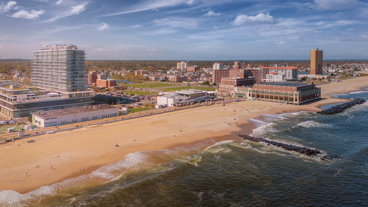 Aerial view of a sandy beach with waves, a pier with buildings, and a cityscape in the background under a partly cloudy sky.