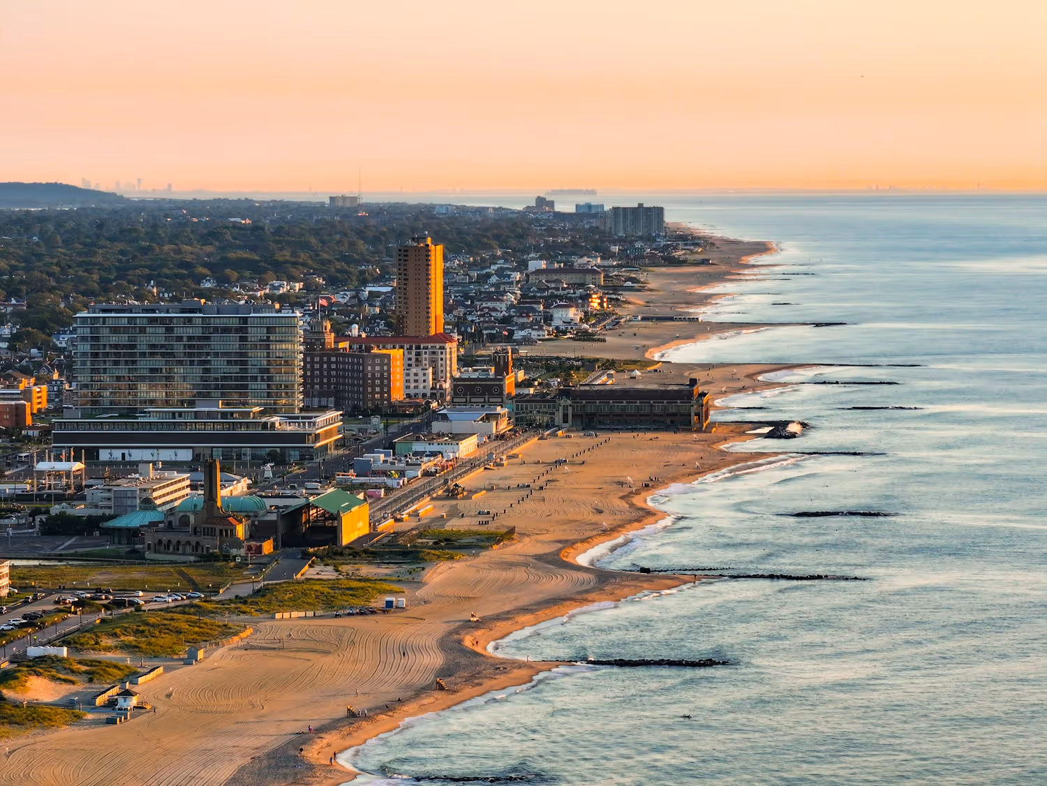 Aerial view of a coastal city at sunset with sandy beaches, buildings, and calm ocean waves.