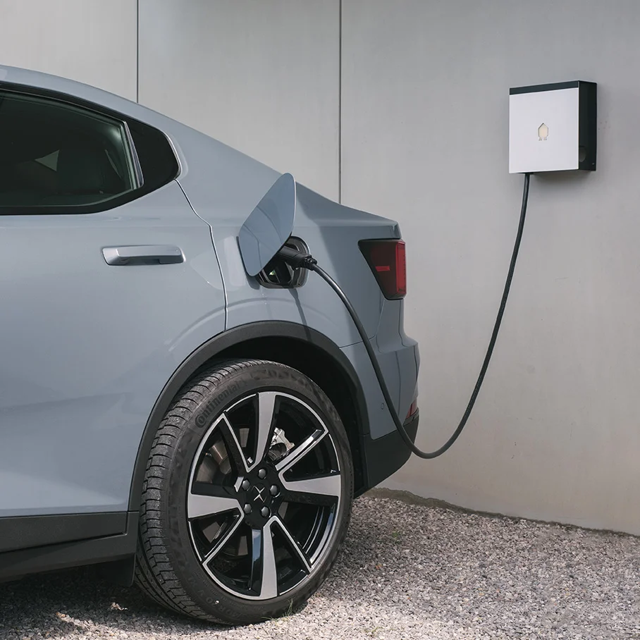Rear side of a gray electric car charging via a wall-mounted charger on a gravel surface.