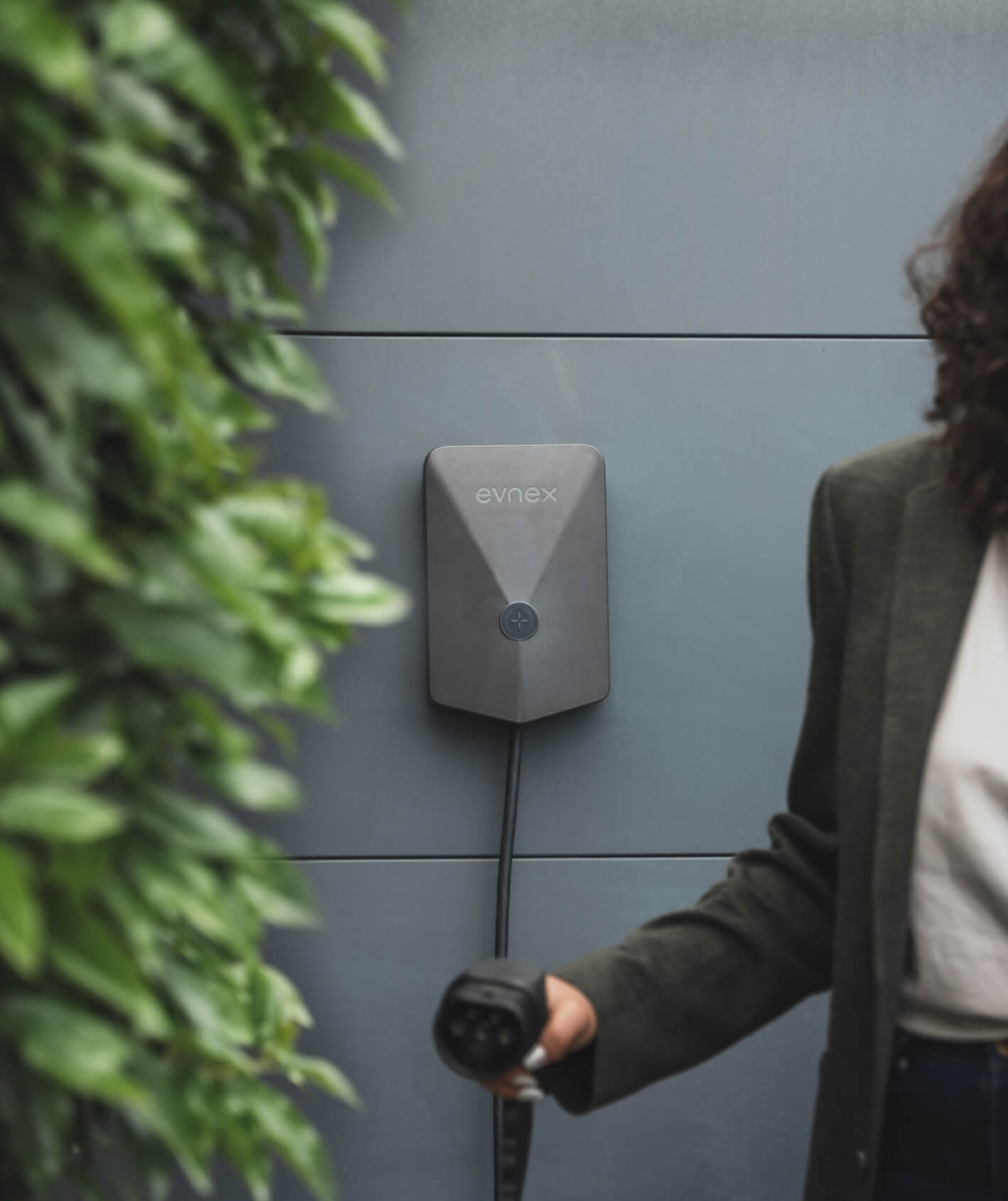 Electric vehicle charging station mounted on a gray wall with a person holding a charging plug nearby.