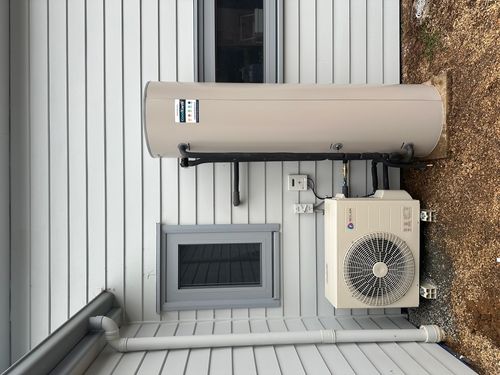 A Reclaim energy heat pump water heater unit installed outside a white house with horizontal siding and two gray-framed windows.