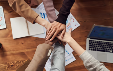 Team members stack their hands together over a desk with charts, notebooks, and a laptop, symbolizing teamwork, collaboration, and shared goals in a workplace setting.