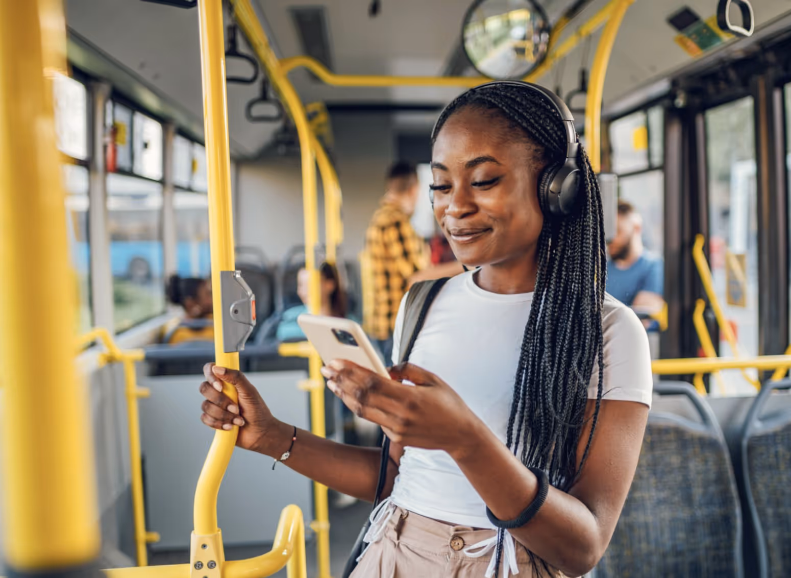 Young woman with braided hair wearing headphones and holding a smartphone while standing and holding a yellow bus pole.