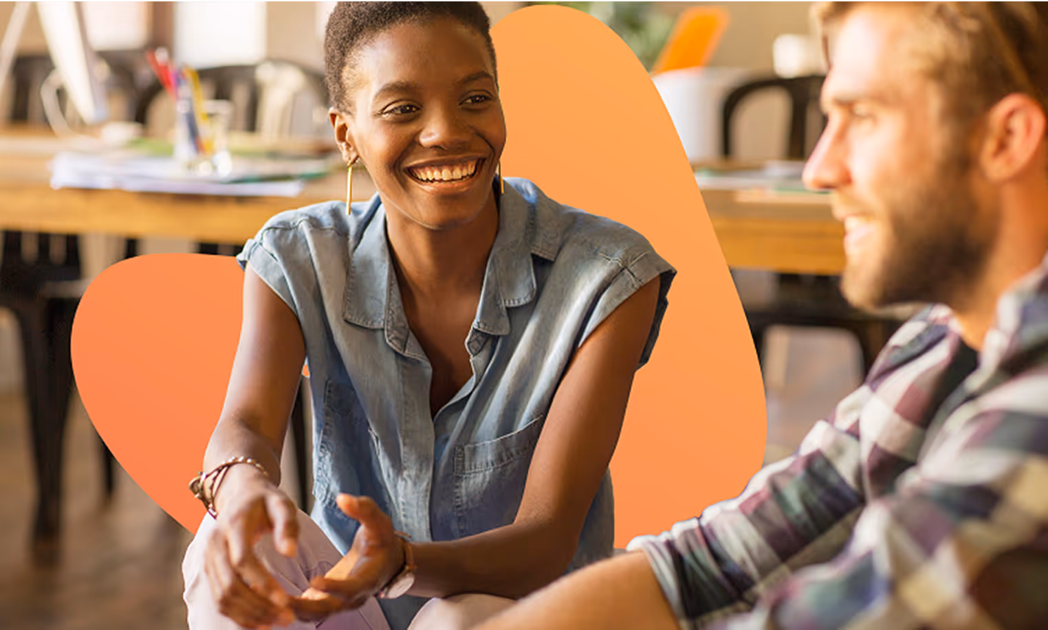 Smiling woman in denim shirt engaged in conversation with a man in a casual indoor setting.