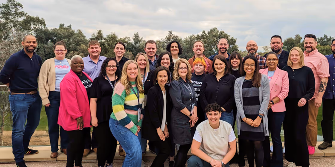 Group of smiling diverse adults posing outdoors with trees and cloudy sky in the background.