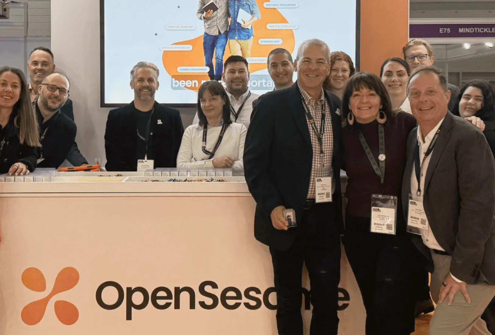 Group of smiling professionals standing behind and in front of a booth with the OpenSesame logo at a conference or event.