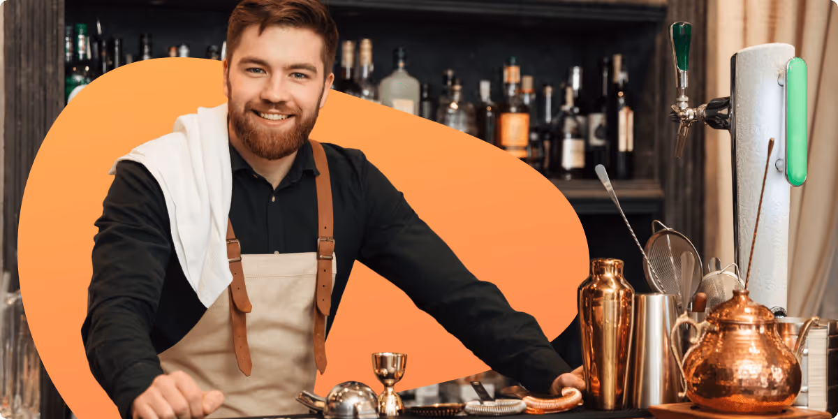 Smiling bearded bartender wearing apron and black shirt standing behind a bar with copper cocktail tools and drink dispenser.