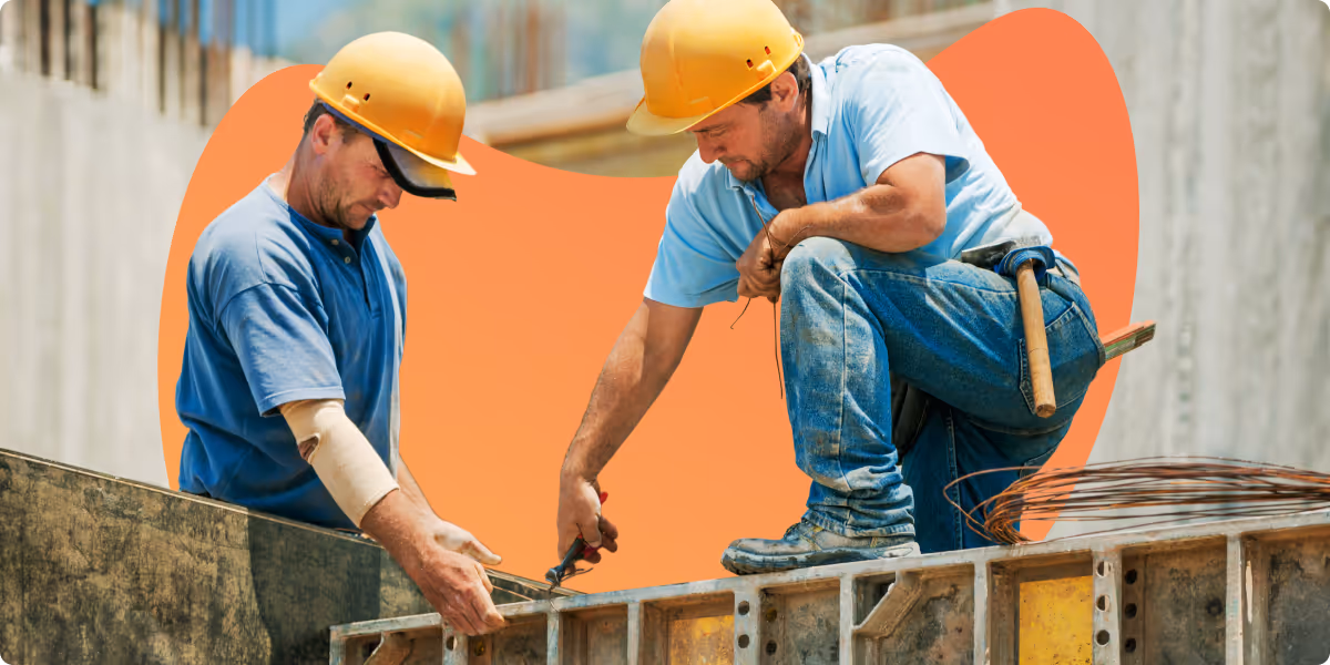 Two construction workers wearing yellow hard hats and blue shirts working on a concrete form.