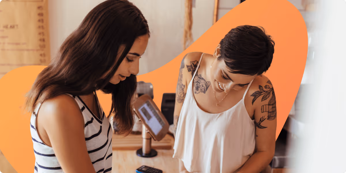 Two women with tattoos and casual sleeveless tops standing and looking down indoors near a payment terminal.