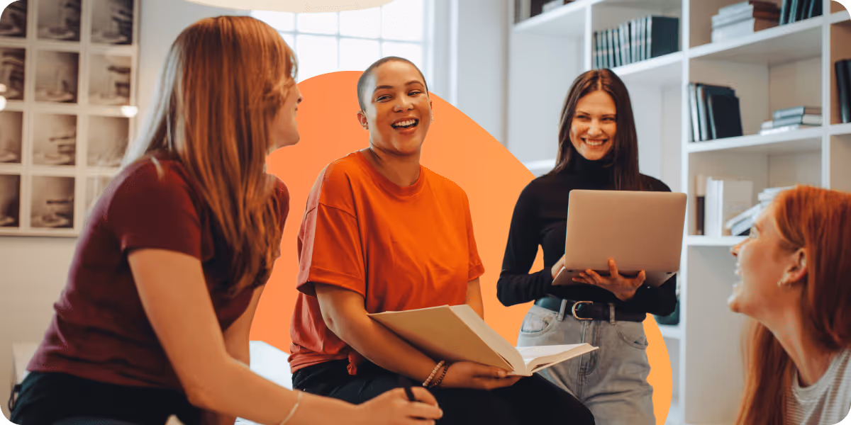 Four women in a casual office setting chatting and smiling, one holding a laptop and another holding an open book.