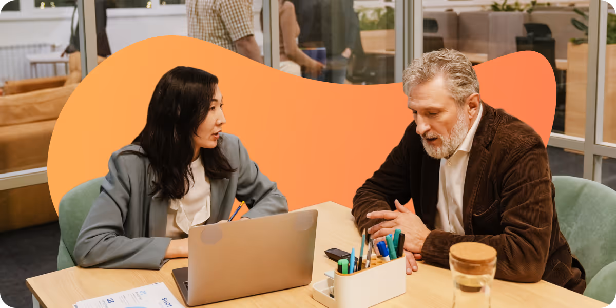 Two colleagues having a discussion at a desk with a laptop, pens, and documents in a modern office setting.