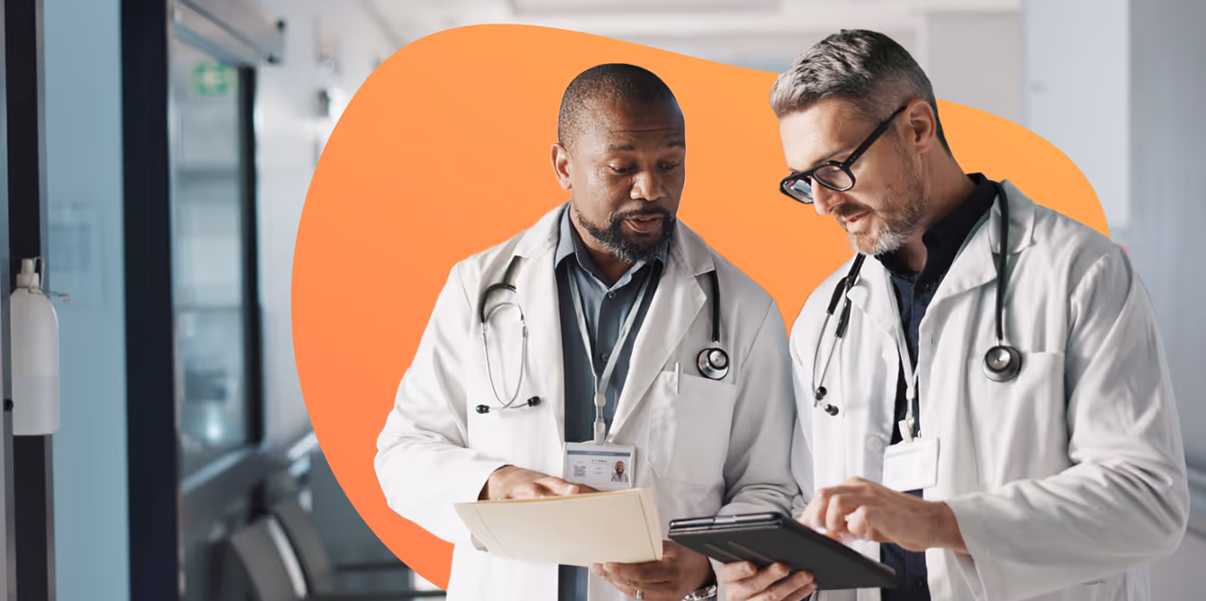 Two doctors wearing white coats and stethoscopes reviewing patient information on a tablet and file in a hospital corridor.