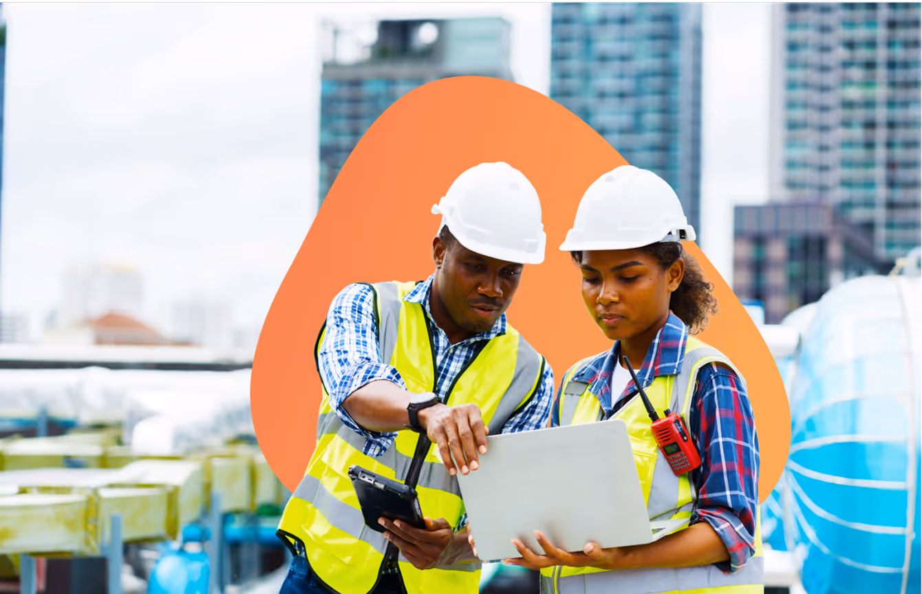 Two construction workers wearing hard hats and reflective vests reviewing information on a laptop and a tablet at an outdoor site.