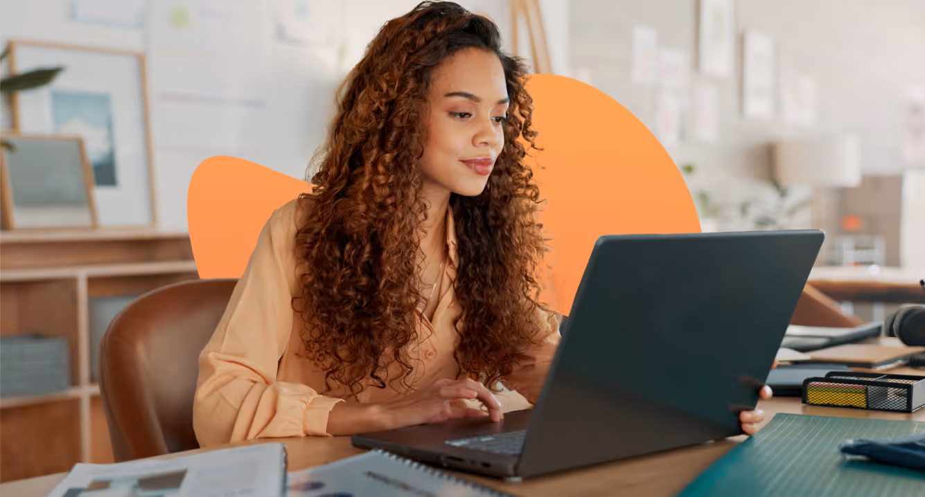 Woman with curly hair working on a laptop at a desk in a modern office setting.