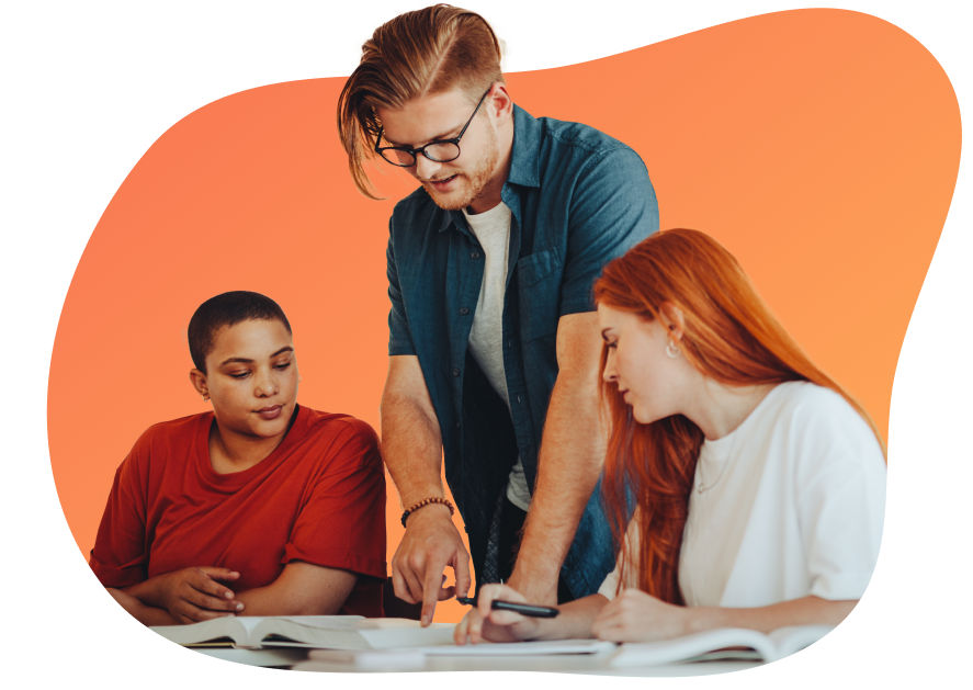 A man with glasses and blue shirt is helping two women with open books, one in a red shirt and the other with long red hair in a white shirt.