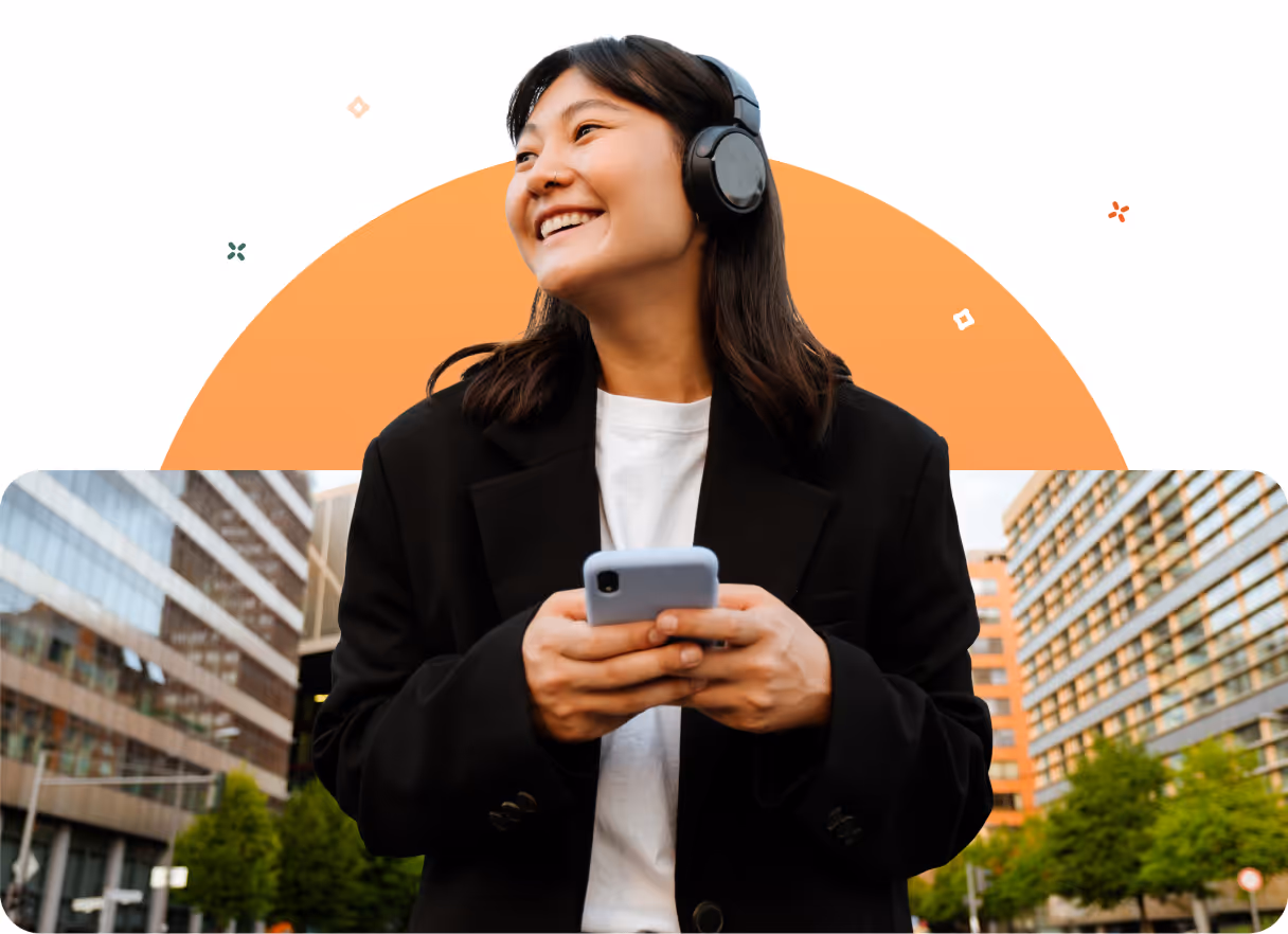 Smiling woman wearing headphones and holding a smartphone outdoors with urban buildings in the background.