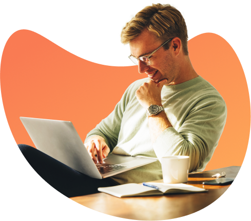 A man sitting at a wooden table, smiling while working on a laptop with a coffee cup and notebook nearby.