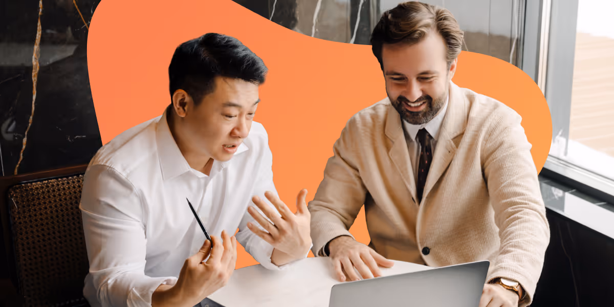 Two men sitting at a table, discussing while looking at a laptop screen, one gesturing with a pen and the other smiling.