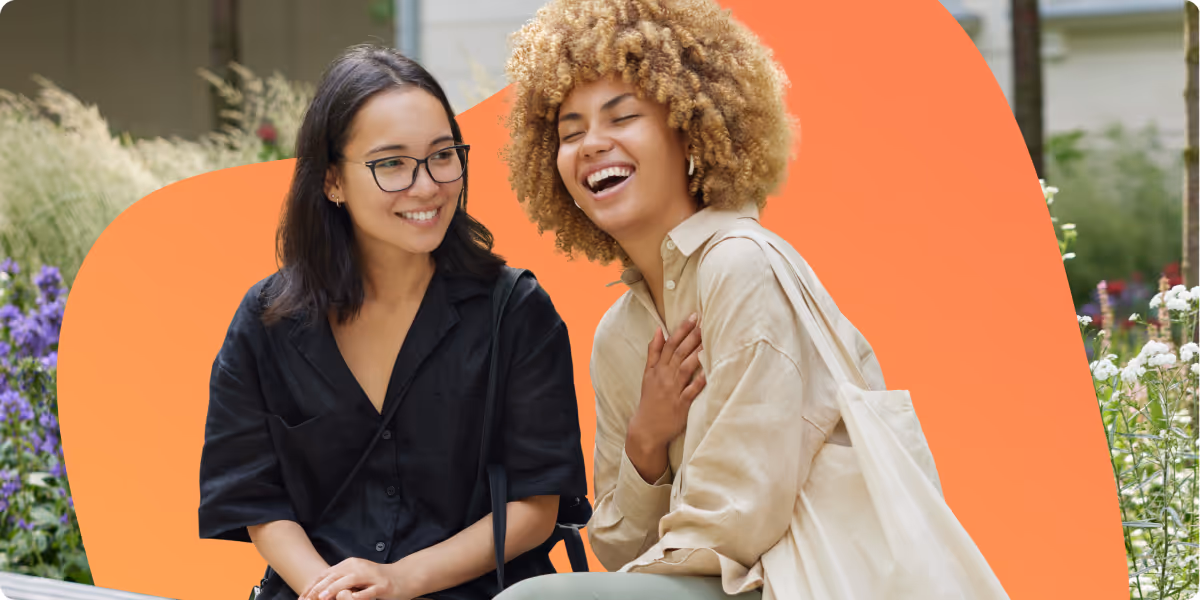 Two young women sitting outdoors, one with glasses smiling and the other laughing with hand on chest.
