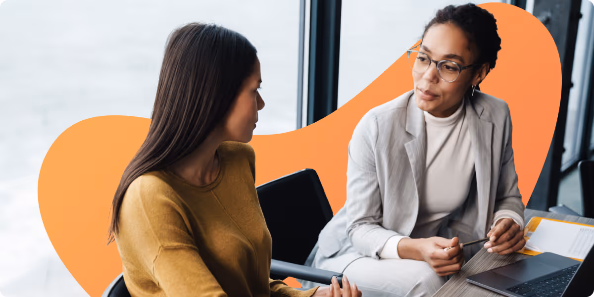Two women having a conversation at a table with a laptop in a bright office setting.